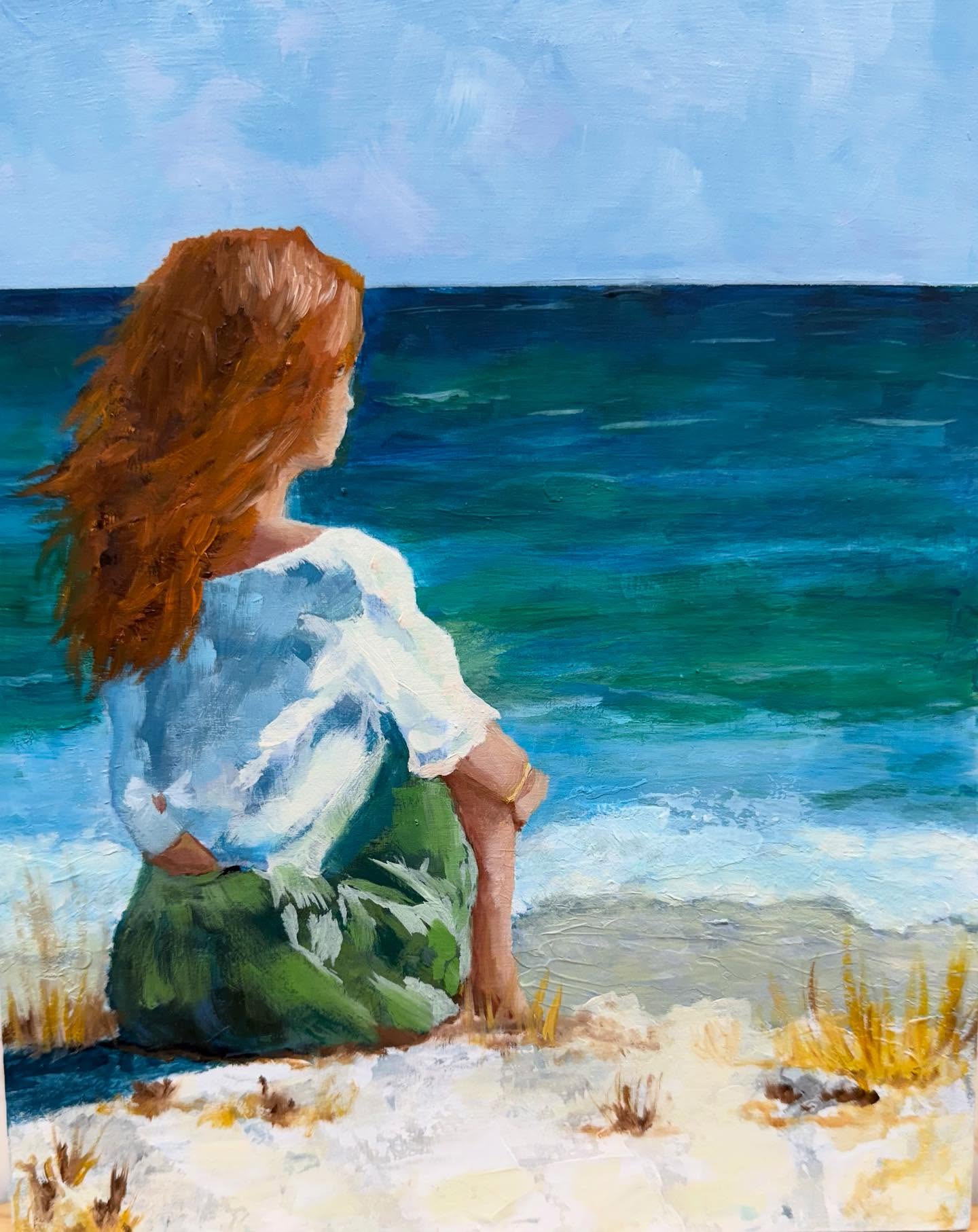 A woman with long,flowing red hair sits looking out over the ocean, knees drawn to her chest.